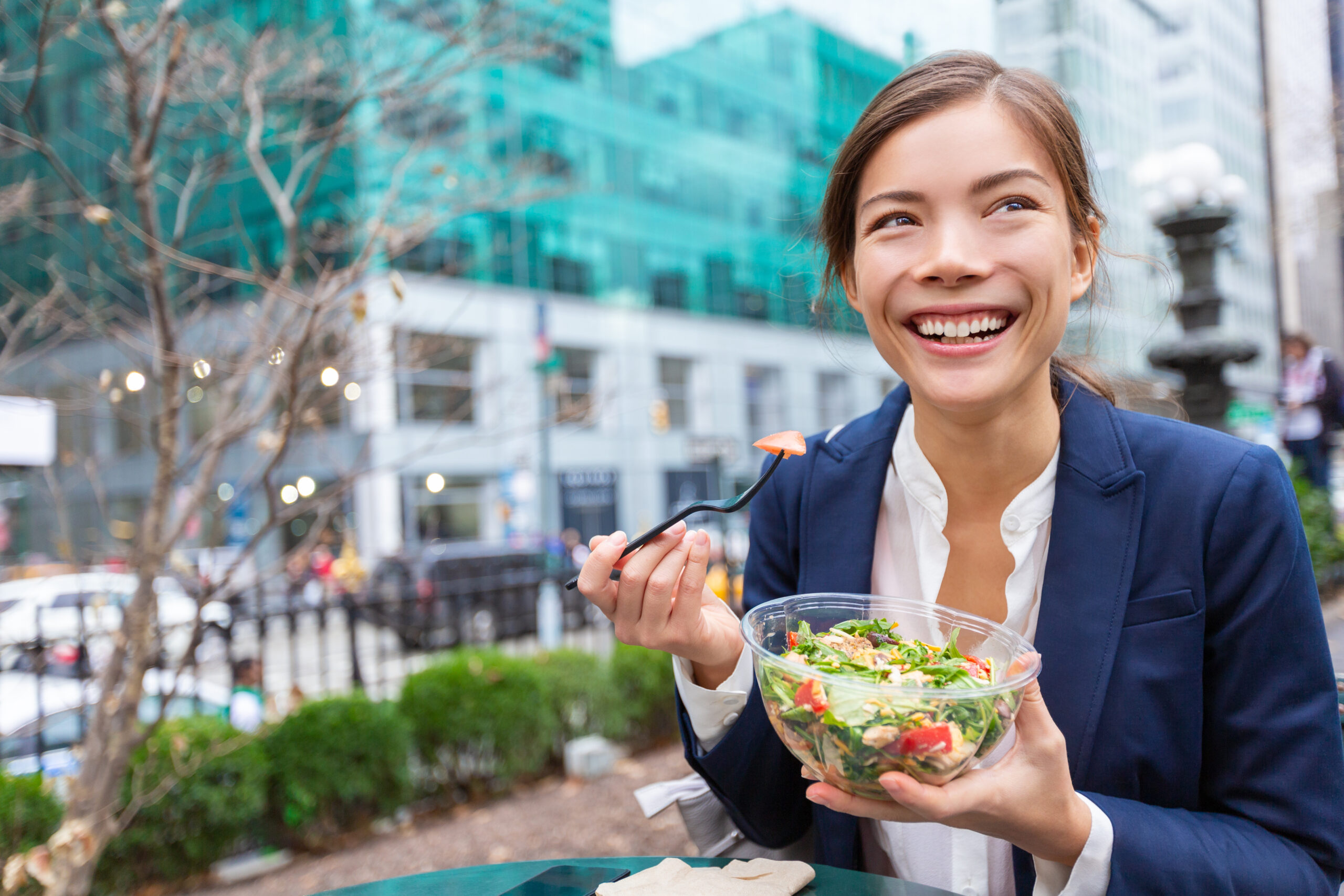 woman in a business suit smiles while taking a bite of salad in an outdoor city setting.